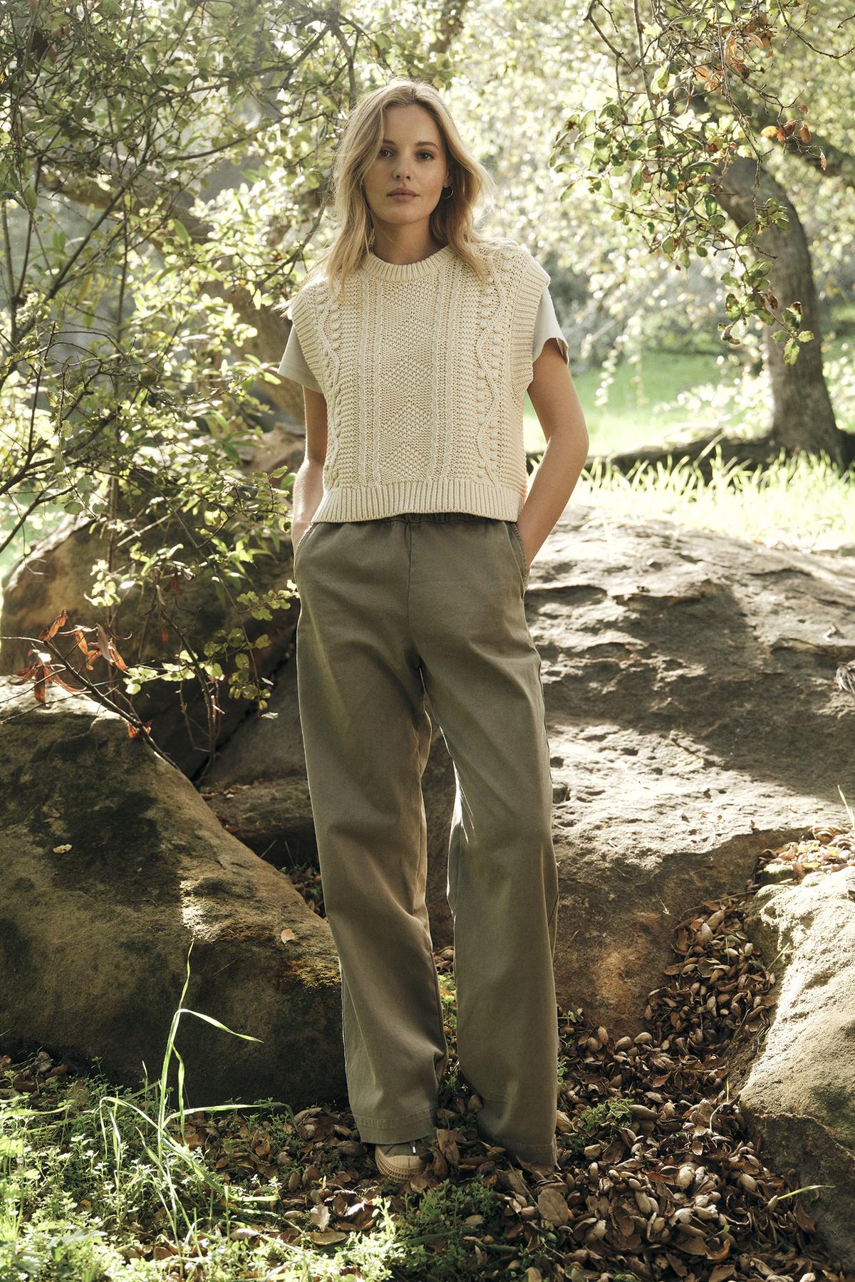   A person standing outdoors on a leafy ground near a large rock. They are wearing the MAGGY SWEATER VEST by Velvet by Graham & Spencer and loose-fitting trousers. Trees and greenery are visible in the background. 