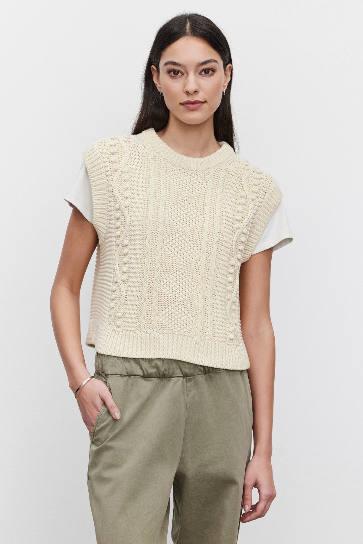   A person with long dark hair is wearing the MAGGY SWEATER VEST in cream from Velvet by Graham & Spencer over a white T-shirt, paired with olive green pants. The background is plain and white. 