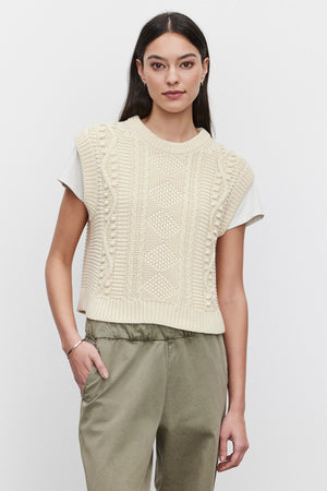 A person with long dark hair is wearing the MAGGY SWEATER VEST in cream from Velvet by Graham & Spencer over a white T-shirt, paired with olive green pants. The background is plain and white.