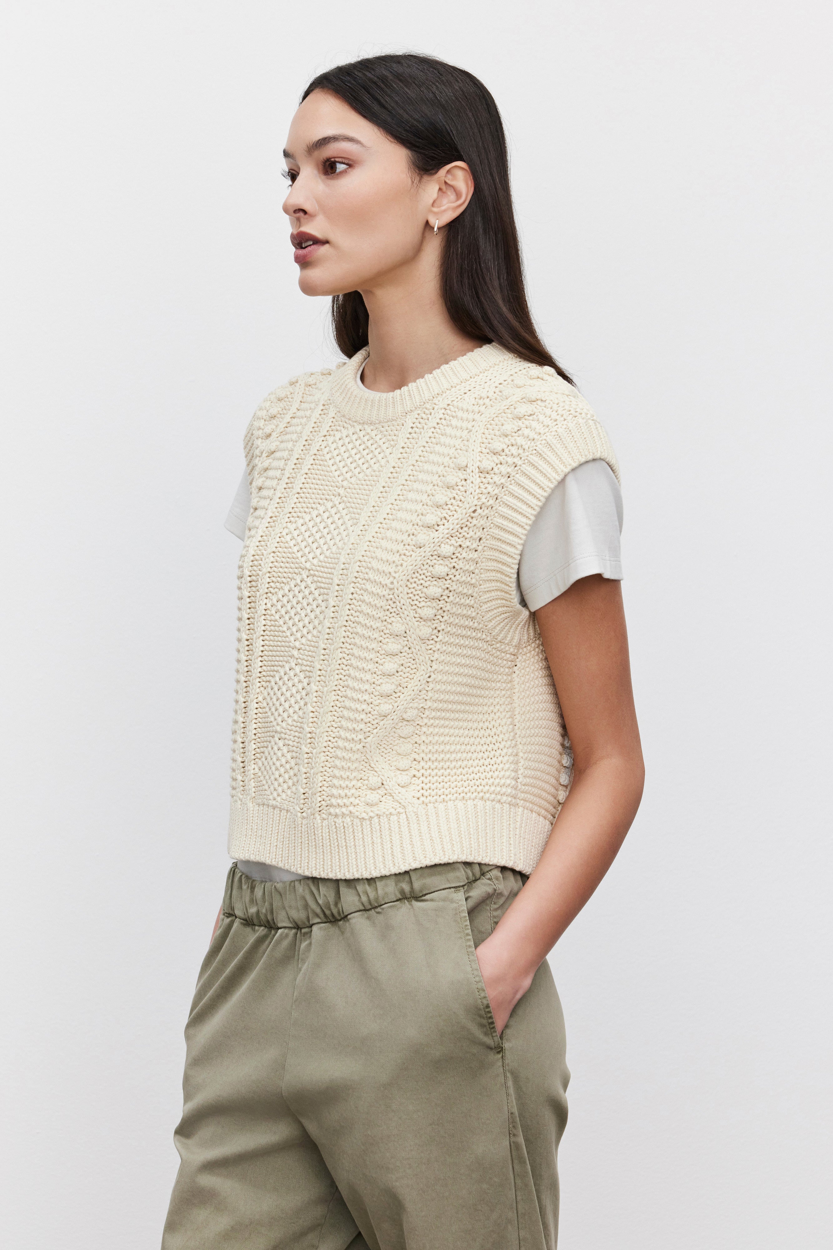   A person stands sideways, donning the MAGGY SWEATER VEST by Velvet by Graham & Spencer in a cream color, layered over a light gray shirt and paired with green pants, with hands in pockets. 