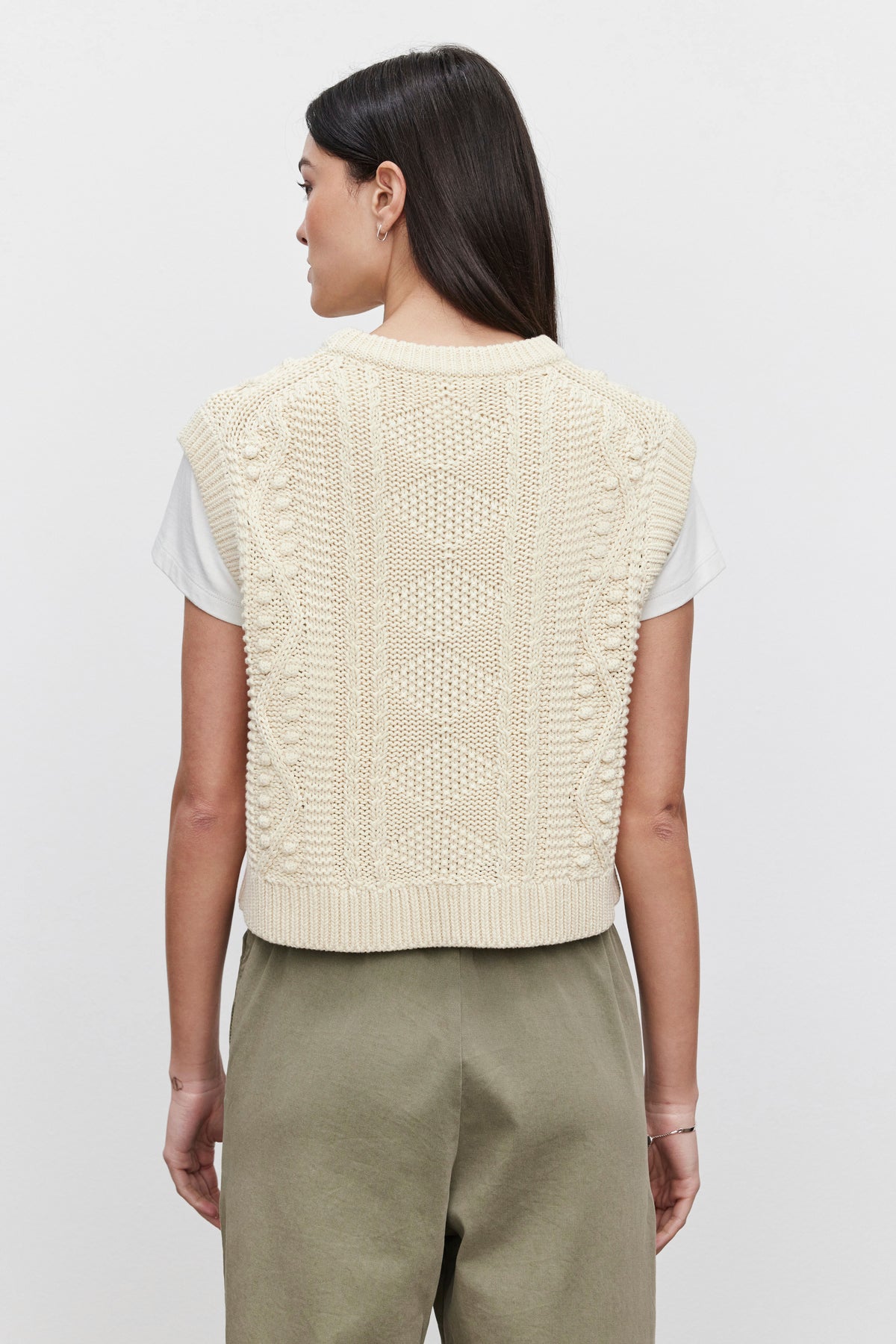   A person with long dark hair is shown from the back, wearing a white shirt under Velvet by Graham & Spencer's MAGGY SWEATER VEST, which features beige cable knit textures, paired with olive green pants. The background is plain and light-colored. 