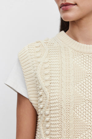 Close-up of a person wearing the MAGGY SWEATER VEST from Velvet by Graham & Spencer, featuring its sleeveless, textured beige design over a white t-shirt. The image highlights the detailed cable knit patterns and focuses on the person's shoulder and part of their face, showcasing the luxurious feel of pima cotton.