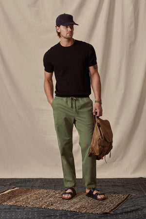 A man in a black t-shirt, green pants, and sandals, wearing a cap and holding a backpack, stands on a Velvet by Graham & Spencer rug against a draped cotton/linen blend canvas background.