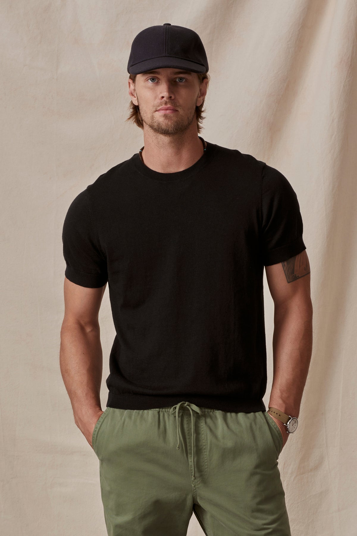   A man wearing a black cotton/linen blend Dexter Crew t-shirt from Velvet by Graham & Spencer and green trousers, accessorized with a cap and a watch, poses against a beige background. 