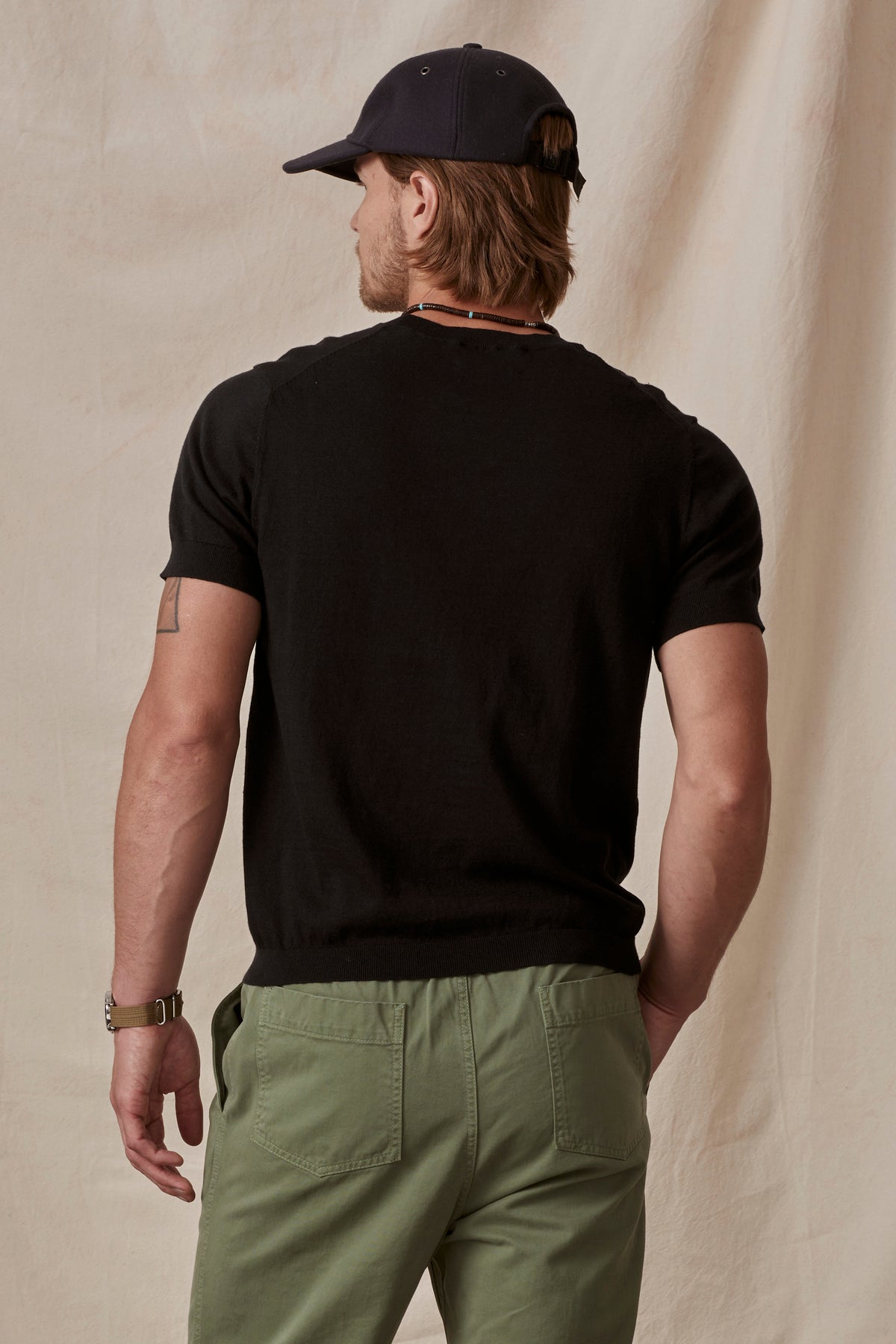   A man viewed from the back, wearing a black cotton/linen blend t-shirt (DEXTER CREW by Velvet by Graham & Spencer) and green pants, with a baseball cap, standing against a beige backdrop. 