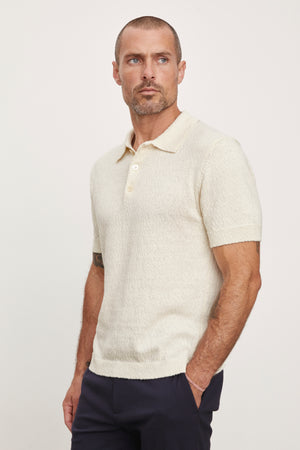 A man with a beard wearing a Velvet by Graham & Spencer Tiberius Polo shirt and dark pants, standing against a light background.