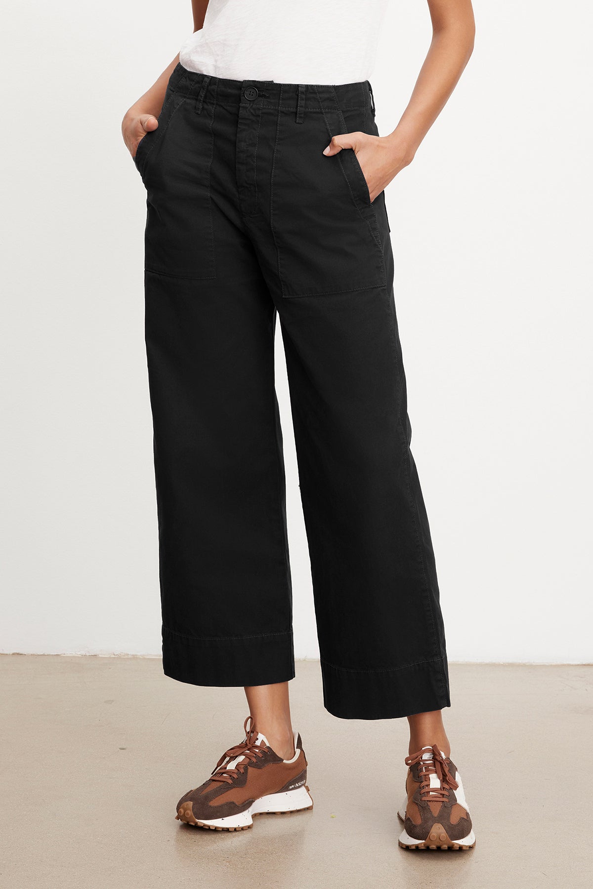   Person wearing black MYA COTTON CANVAS PANT by Velvet by Graham & Spencer with a flared leg and brown sneakers with white soles, standing against a plain white background. Hands are in the pants pockets. 