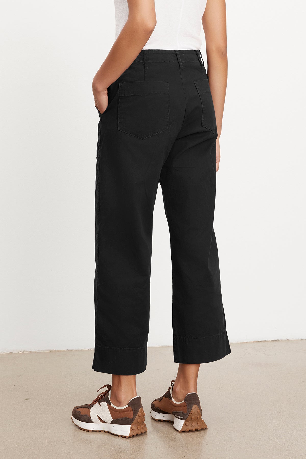 A person wearing black MYA COTTON CANVAS PANTS by Velvet by Graham & Spencer, a white slim top, and brown sneakers stands facing away, with one hand in their pocket.-36409563381953