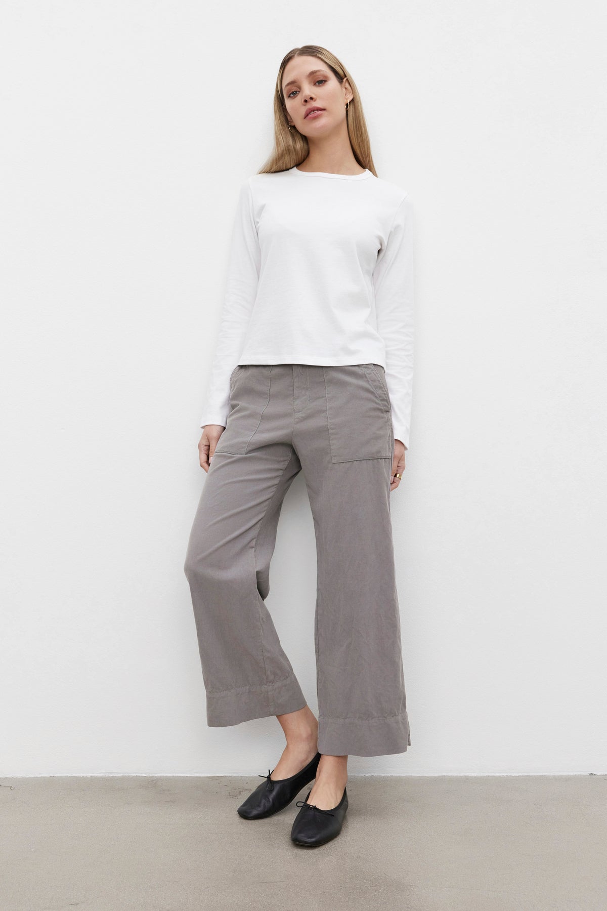   A person stands against a plain white wall wearing a white long-sleeve shirt, flattering VERA CORDUROY PANT by Velvet by Graham & Spencer, and black shoes. 