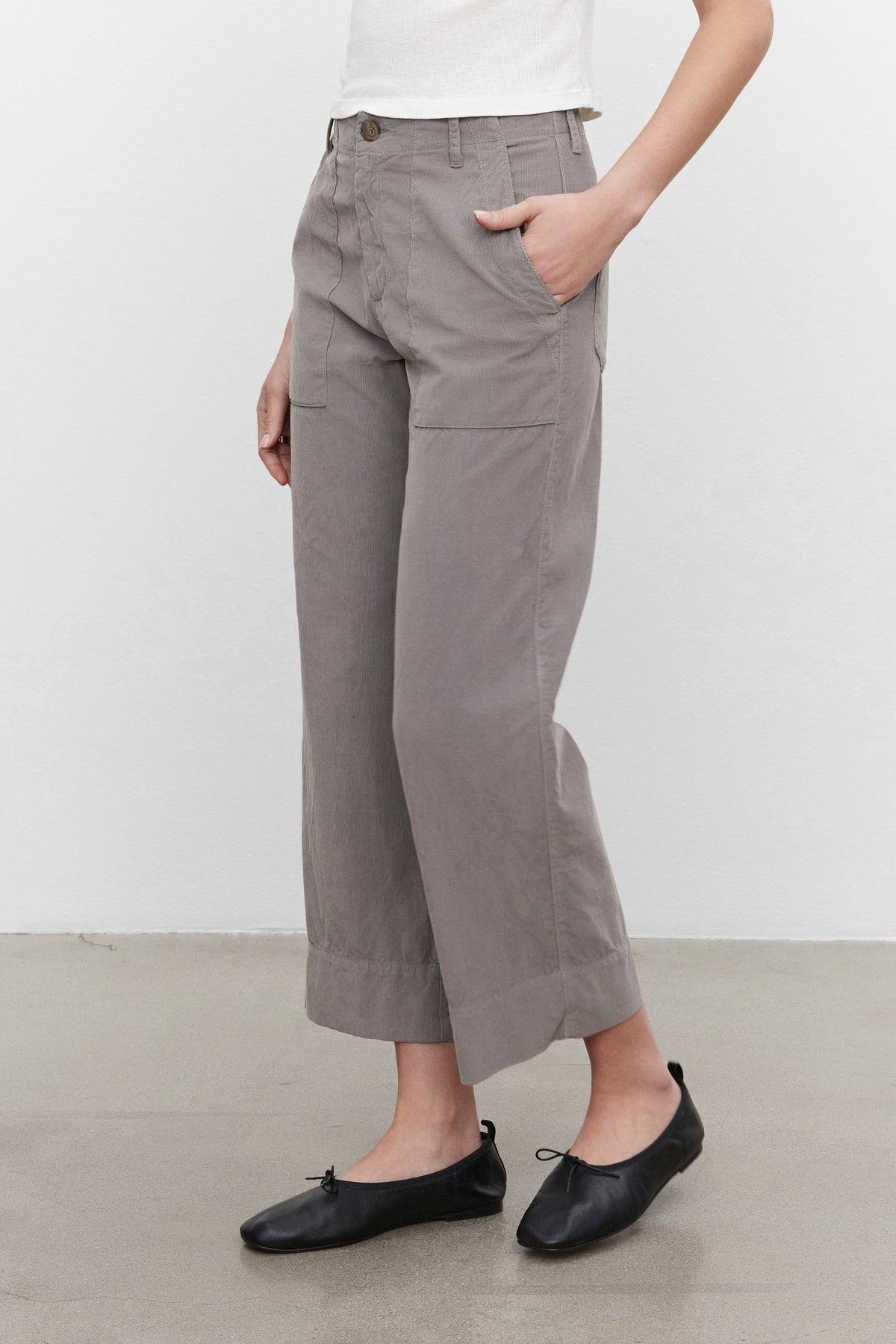   A person stands on a concrete floor, sporting a white top, VERA CORDUROY PANT in gray pinwale cropped wide-leg style by Velvet by Graham & Spencer, and black ballet flats, with one hand casually tucked into a pants pocket. 