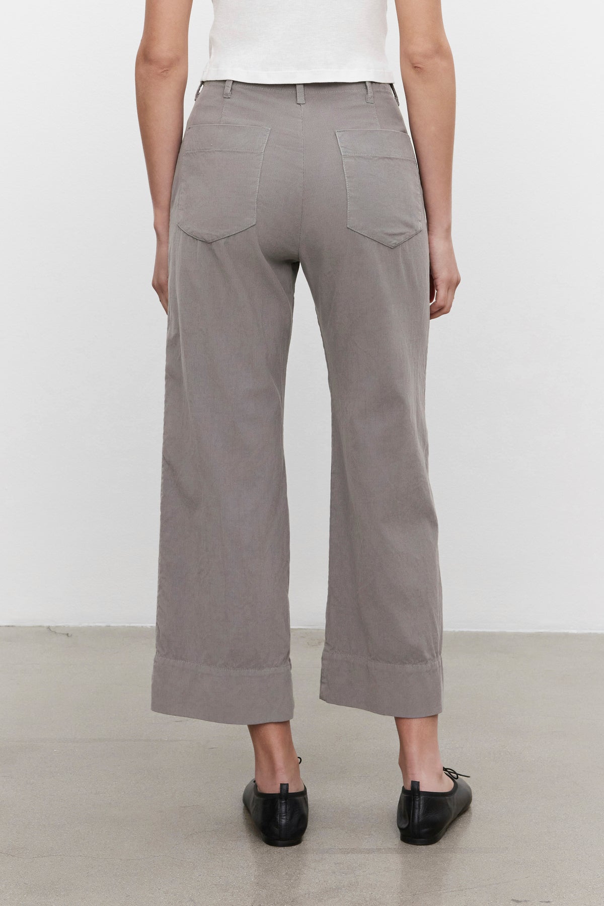   A person stands with their back to the camera, donning the VERA CORDUROY PANT in light gray by Velvet by Graham & Spencer, which creates a flattering silhouette. They pair it with black shoes against a plain background. 