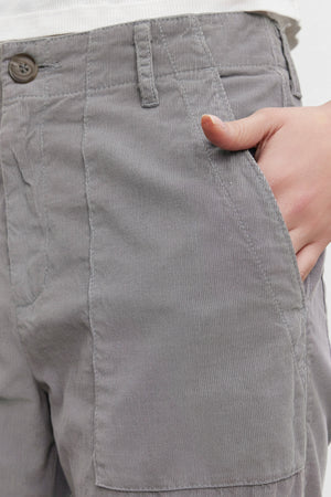 Close-up of a person wearing the VERA CORDUROY PANT by Velvet by Graham & Spencer in gray pinwale corduroy, with their right hand in the pocket. The pants feature a visible button and zipper closure, providing a flattering silhouette.