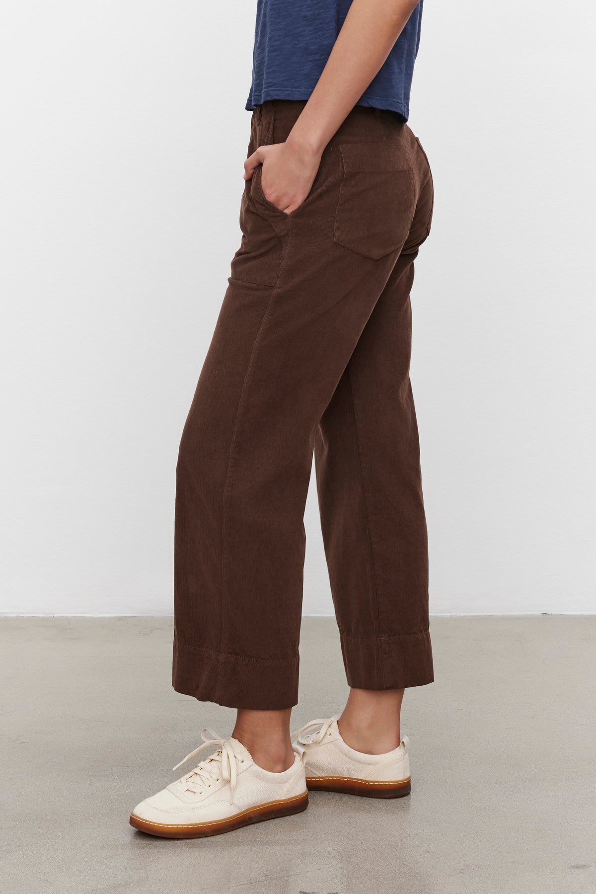   A person wearing the VERA CORDUROY PANT from Velvet by Graham & Spencer, complemented by a blue shirt and cream-colored sneakers, stands on a grey concrete floor with a white wall in the background. The individual is in a relaxed pose with hands in pockets. 