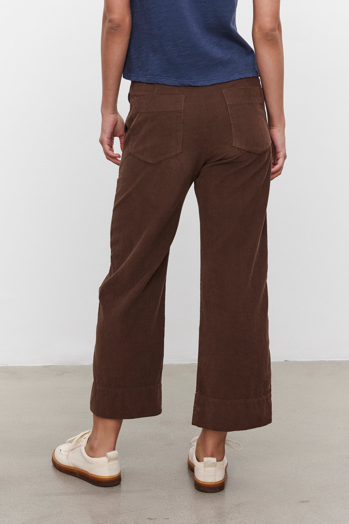   A person wearing a blue shirt and the VERA CORDUROY PANT by Velvet by Graham & Spencer is standing on a gray floor facing away from the camera. 