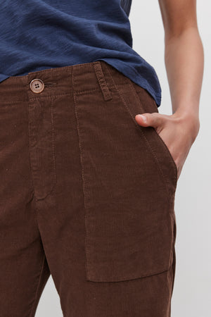 A person wearing a blue shirt and Velvet by Graham & Spencer's VERA CORDUROY PANT in brown, with one hand in their pocket.