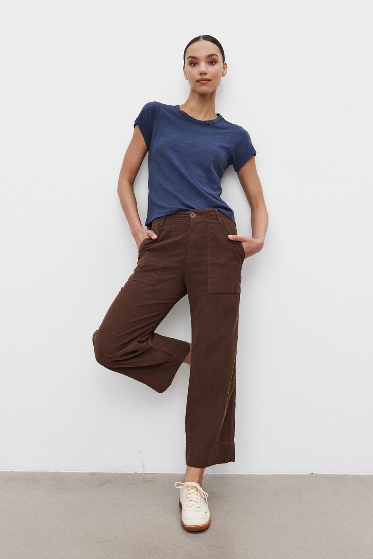   A person stands with hands in pockets, wearing a blue t-shirt, Velvet by Graham & Spencer's VERA CORDUROY PANT in pinwale corduroy cropped wide-leg style, and white sneakers. The backdrop is a plain white wall. 