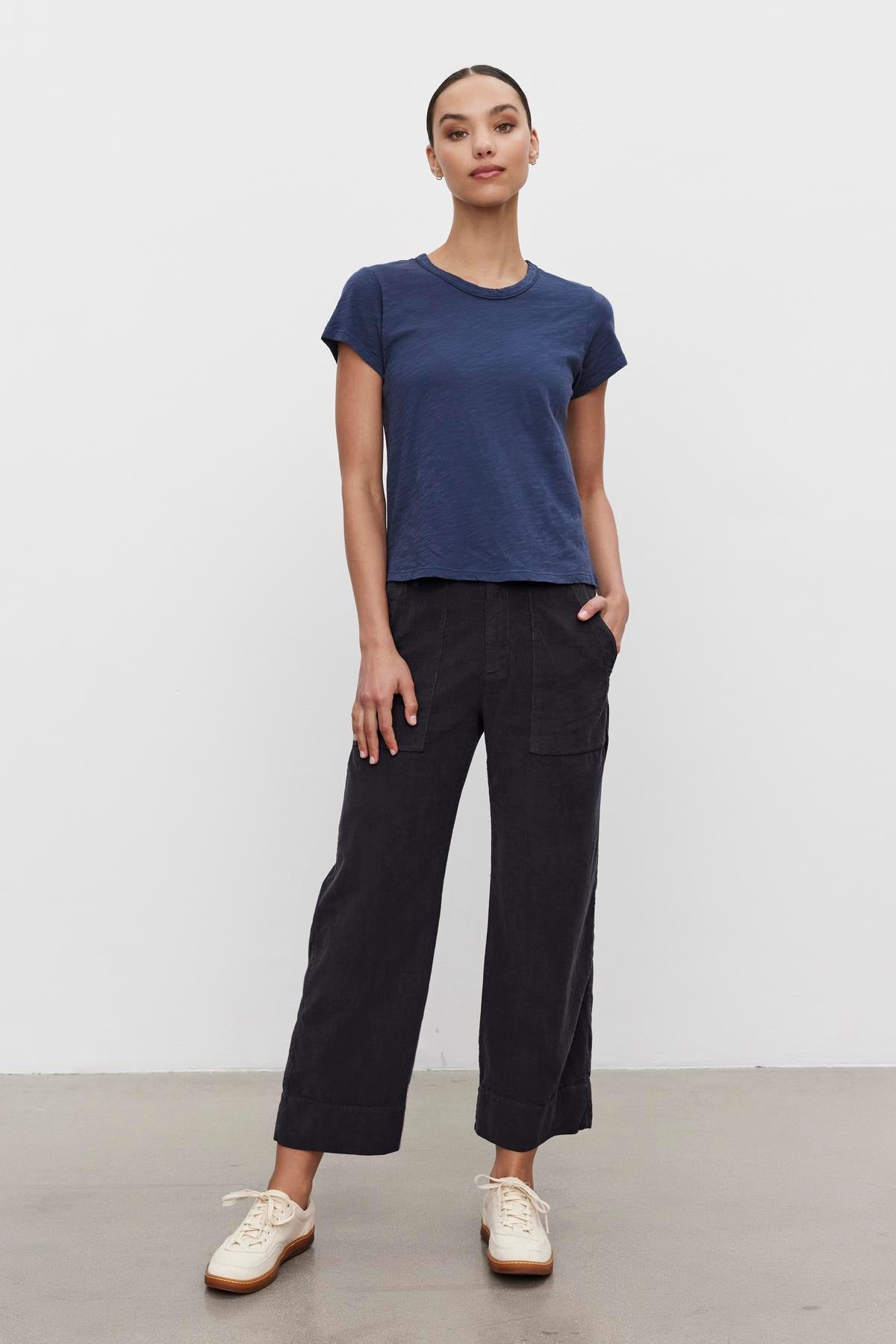 Person standing against a plain white background, wearing a navy T-shirt, Velvet by Graham & Spencer VERA CORDUROY PANT in black, and beige sneakers. One hand is in their pocket, and they are looking at the camera.-37665605517505