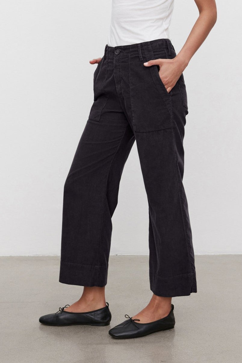   A person is standing against a plain backdrop wearing a white tank top, Velvet by Graham & Spencer's VERA CORDUROY PANT in black, and black flats. Hands are in the pockets, looking down at the ground. 