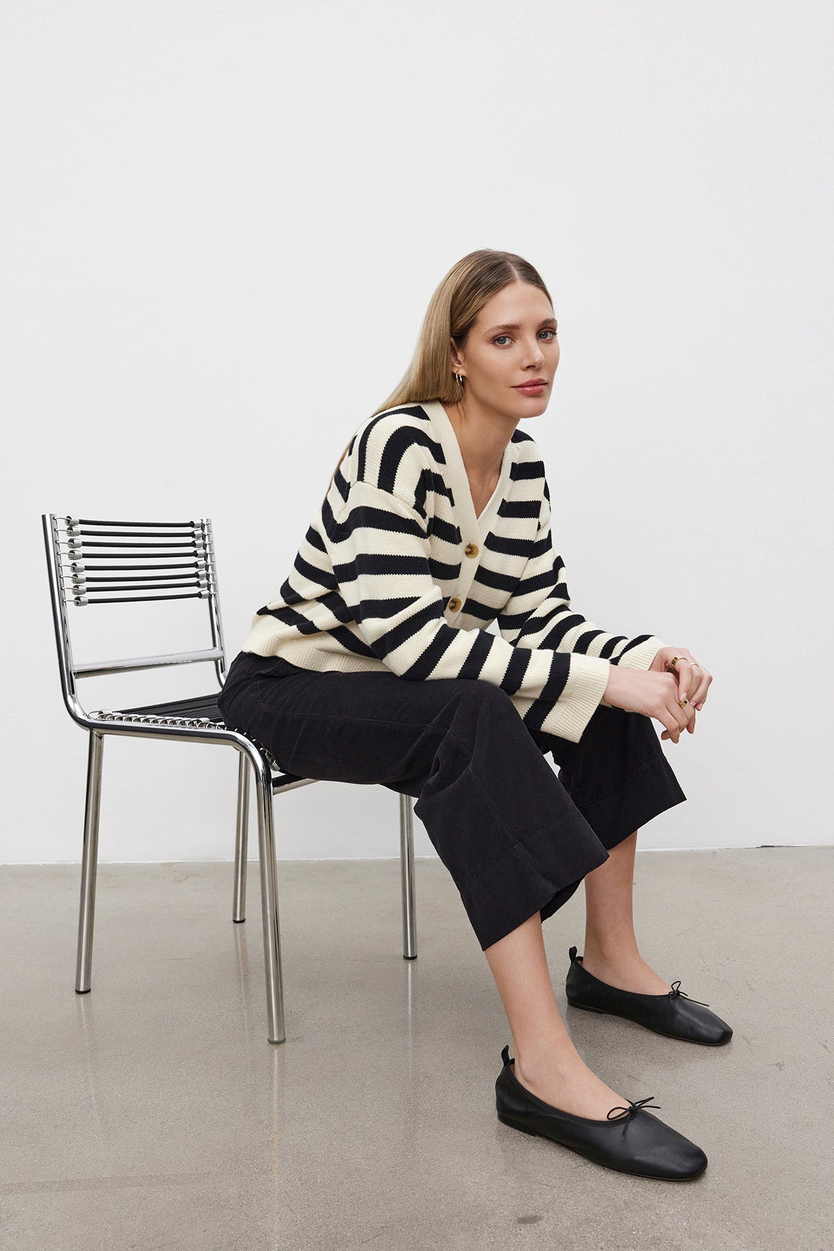 A person wearing a striped sweater, VERA CORDUROY PANT by Velvet by Graham & Spencer, and black shoes sits on a metal chair in front of a white wall.-38426459603137