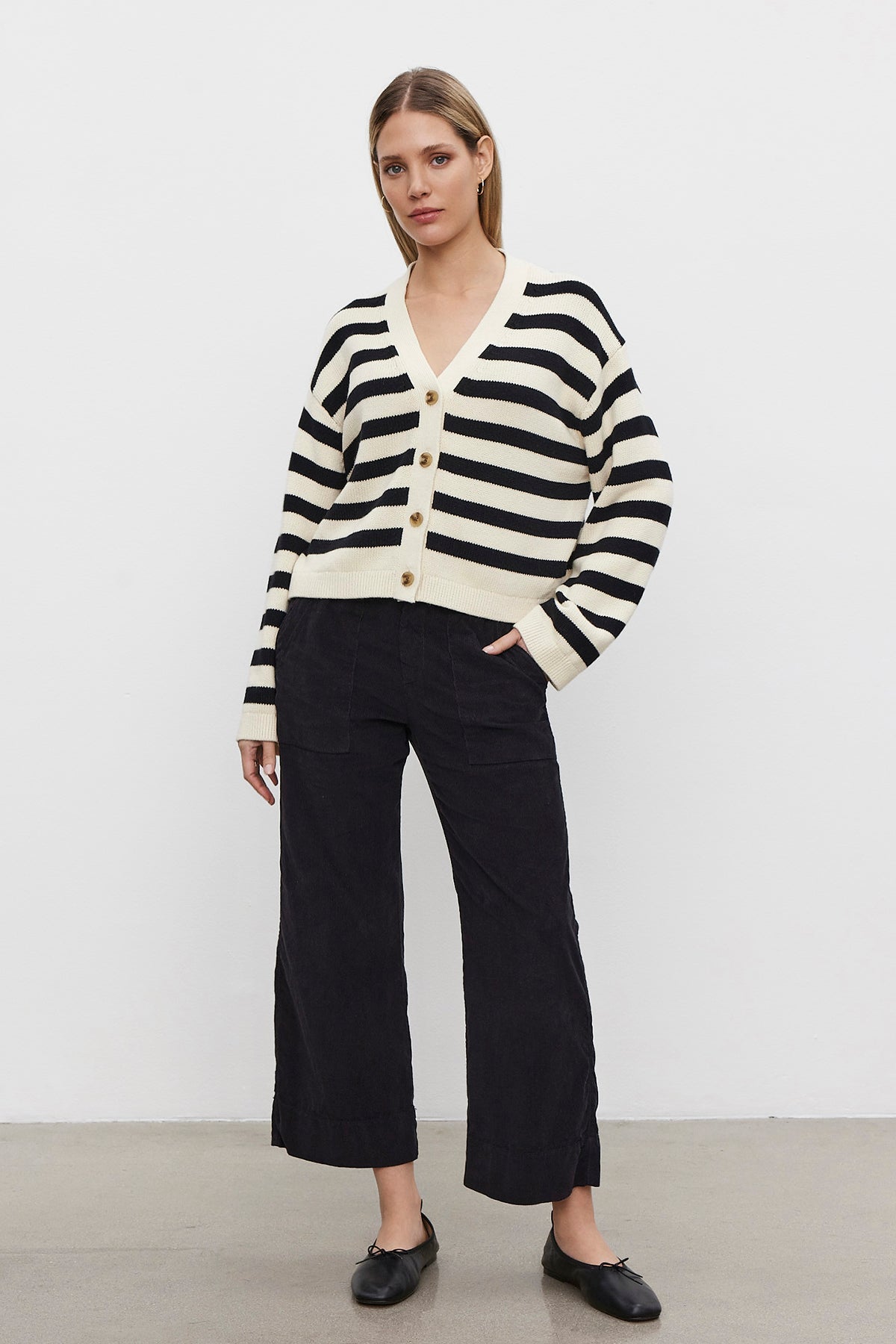   A person stands on a gray floor with a plain white background, donning a black and white striped cardigan. They're wearing the chic VERA CORDUROY PANT by Velvet by Graham & Spencer, with subtle utility details and sleek black shoes, effortlessly blending style and function. 