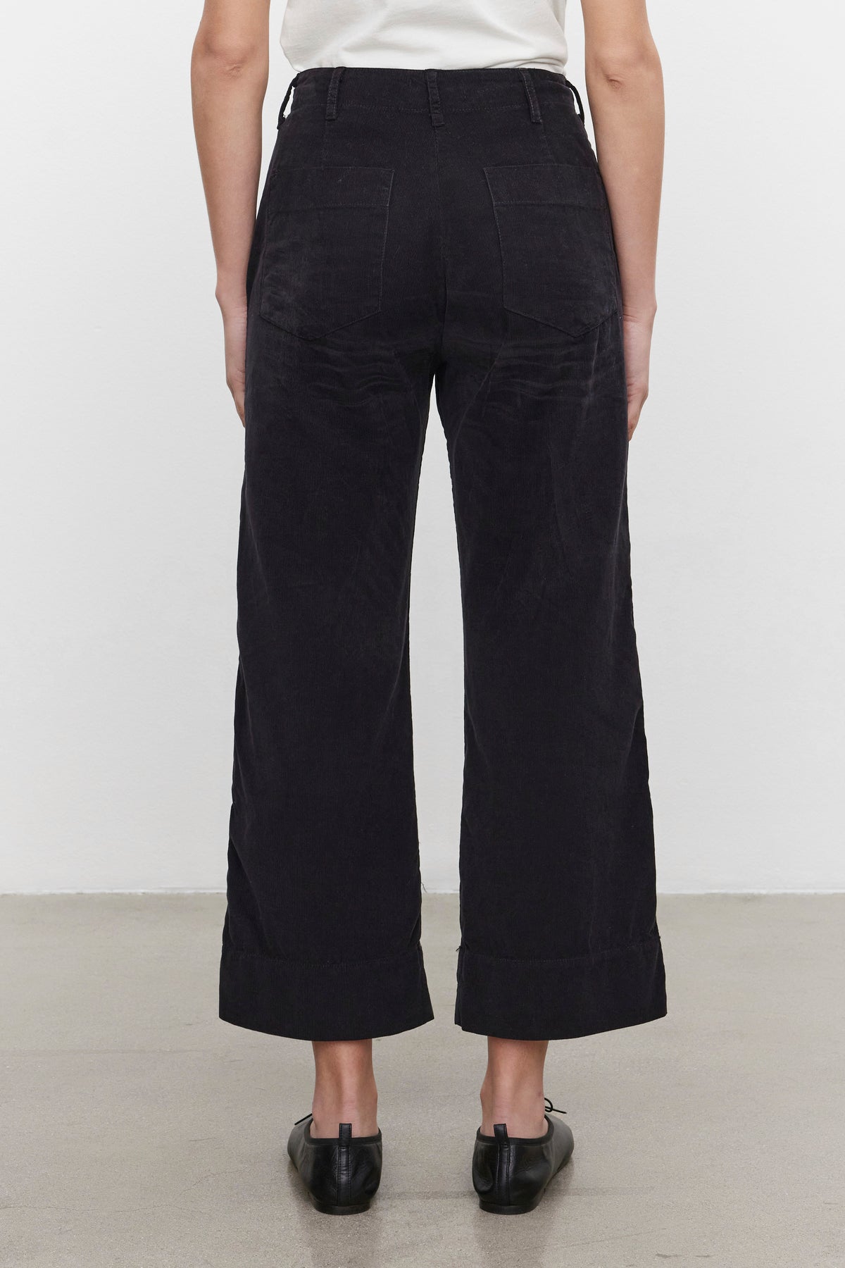 Back view of a person wearing the Velvet by Graham & Spencer VERA CORDUROY PANT in black pinwale corduroy, paired with a white top and black shoes, standing on a grey floor against a plain white background.-37665605615809