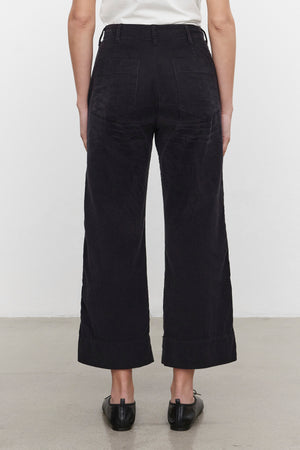 Back view of a person wearing the Velvet by Graham & Spencer VERA CORDUROY PANT in black pinwale corduroy, paired with a white top and black shoes, standing on a grey floor against a plain white background.