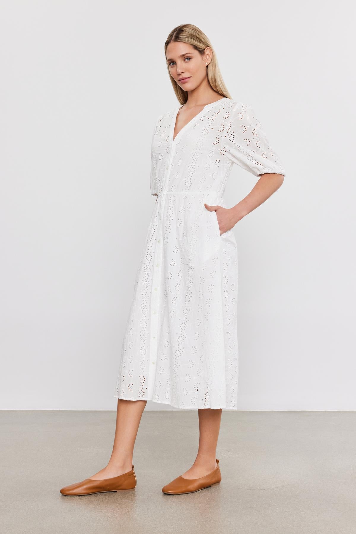   Woman in a white cotton eyelet RORI DRESS with puff sleeves and brown flat shoes, standing against a plain gray background from Velvet by Graham & Spencer. 