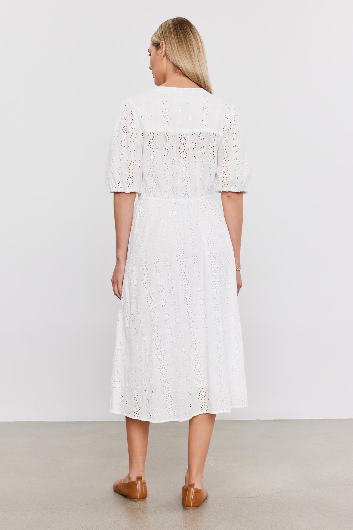 Woman in a white cotton eyelet mid-length RORI DRESS with puff sleeves, viewed from behind, standing against a plain background. She's wearing brown loafers by Velvet by Graham & Spencer.-36910267990209