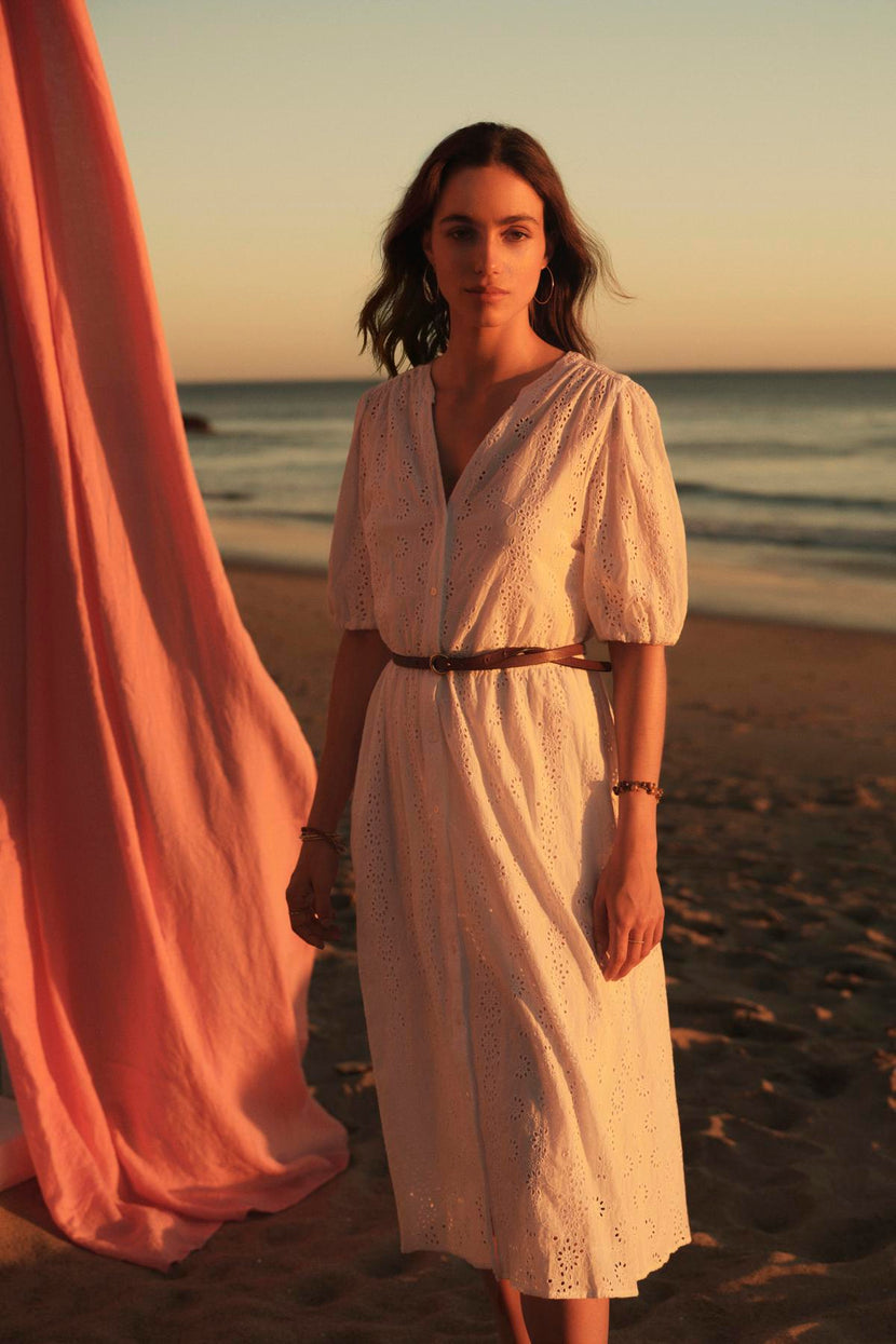 A woman in a white cotton eyelet RORI DRESS from Velvet by Graham & Spencer stands on a beach at sunset, with a pink fabric billowing in the background.