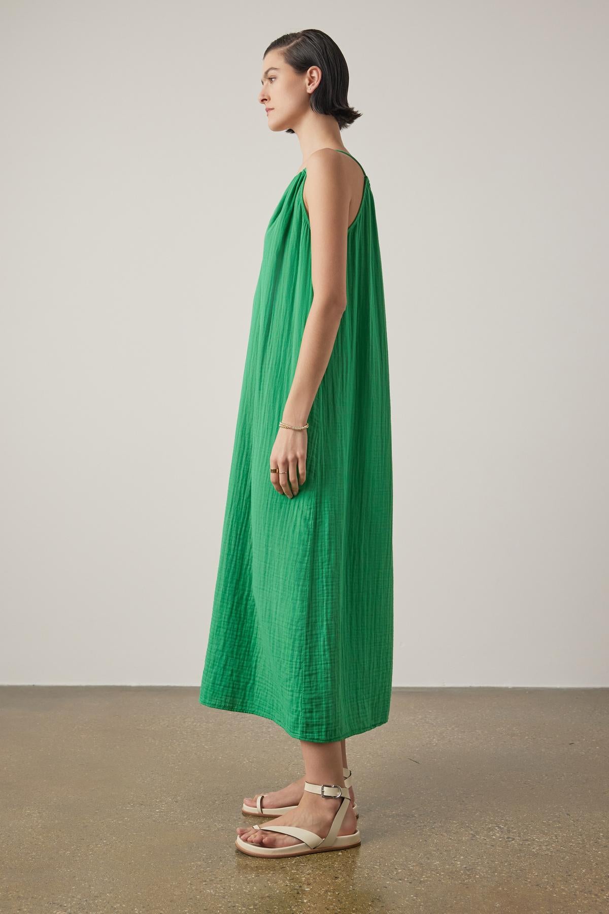   A woman standing in profile, wearing a long green sleeveless Carrillo dress by Velvet by Jenny Graham and beige sandals, against a neutral background. 