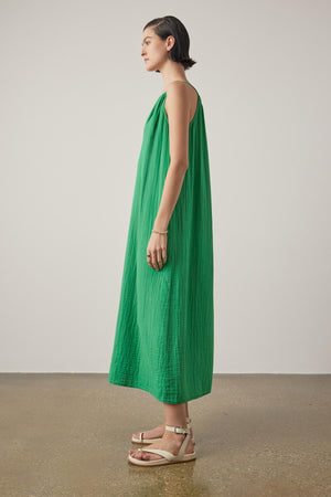 A woman standing in profile, wearing a long green sleeveless Carrillo dress by Velvet by Jenny Graham and beige sandals, against a neutral background.