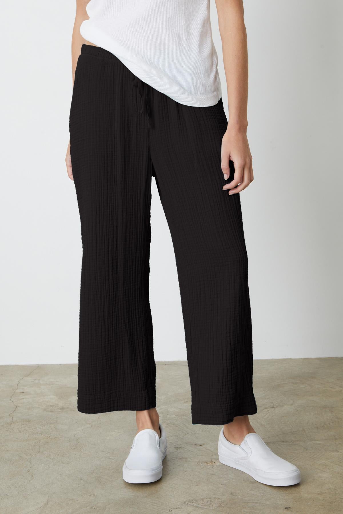 Person wearing black FRANNY COTTON GAUZE PANT by Velvet by Graham & Spencer, a white t-shirt, and white slip-on shoes, standing on a concrete floor against a plain white backdrop.-37238901899457