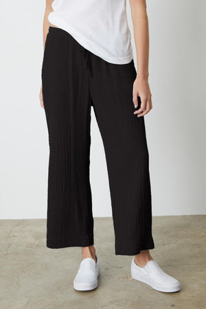 Person wearing black FRANNY COTTON GAUZE PANT by Velvet by Graham & Spencer, a white t-shirt, and white slip-on shoes, standing on a concrete floor against a plain white backdrop.