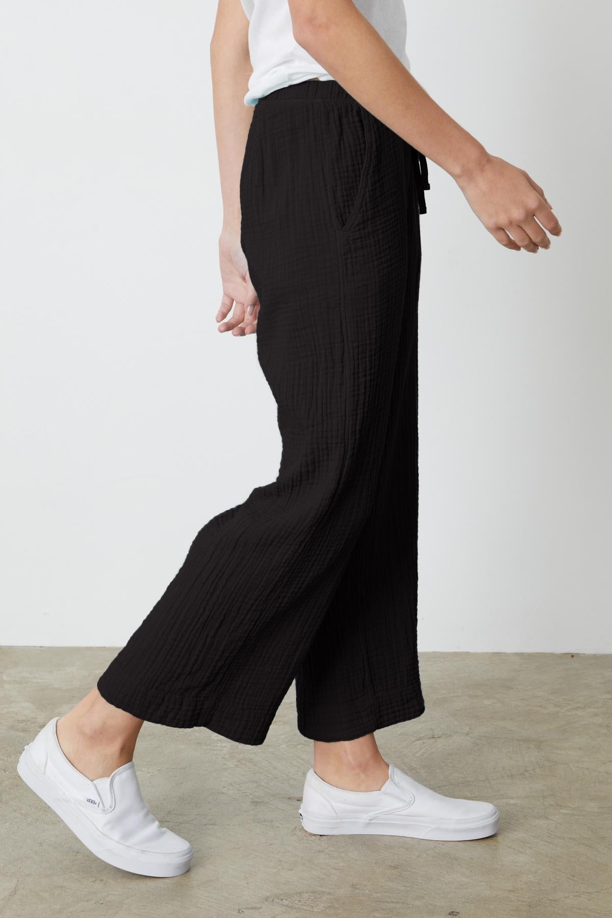   Person wearing black FRANNY COTTON GAUZE PANT by Velvet by Graham & Spencer with a relaxed leg and white slip-on shoes is shown from the side against a plain background. 