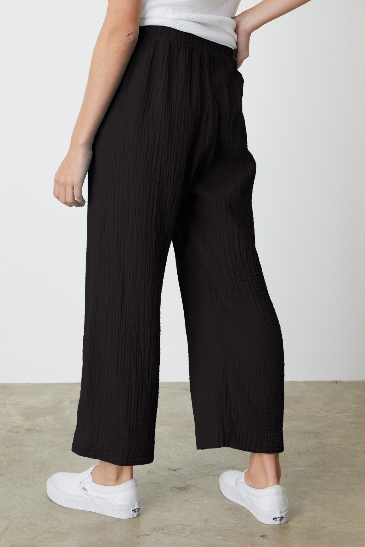   Person wearing black, loose-fitting FRANNY COTTON GAUZE PANT by Velvet by Graham & Spencer with a relaxed leg and a white top, standing sideways with one hand on hip. They are also wearing white sneakers. 