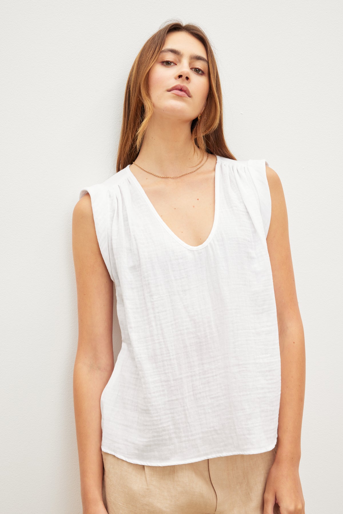   A person with long hair is wearing the Velvet by Graham & Spencer JAYLA COTTON GAUZE TANK TOP, a sleeveless white V-neck top with subtle shirred detailing, and beige pants, standing against a plain white background. 