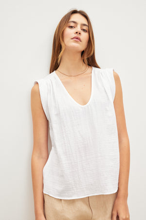 A person with long hair is wearing the Velvet by Graham & Spencer JAYLA COTTON GAUZE TANK TOP, a sleeveless white V-neck top with subtle shirred detailing, and beige pants, standing against a plain white background.