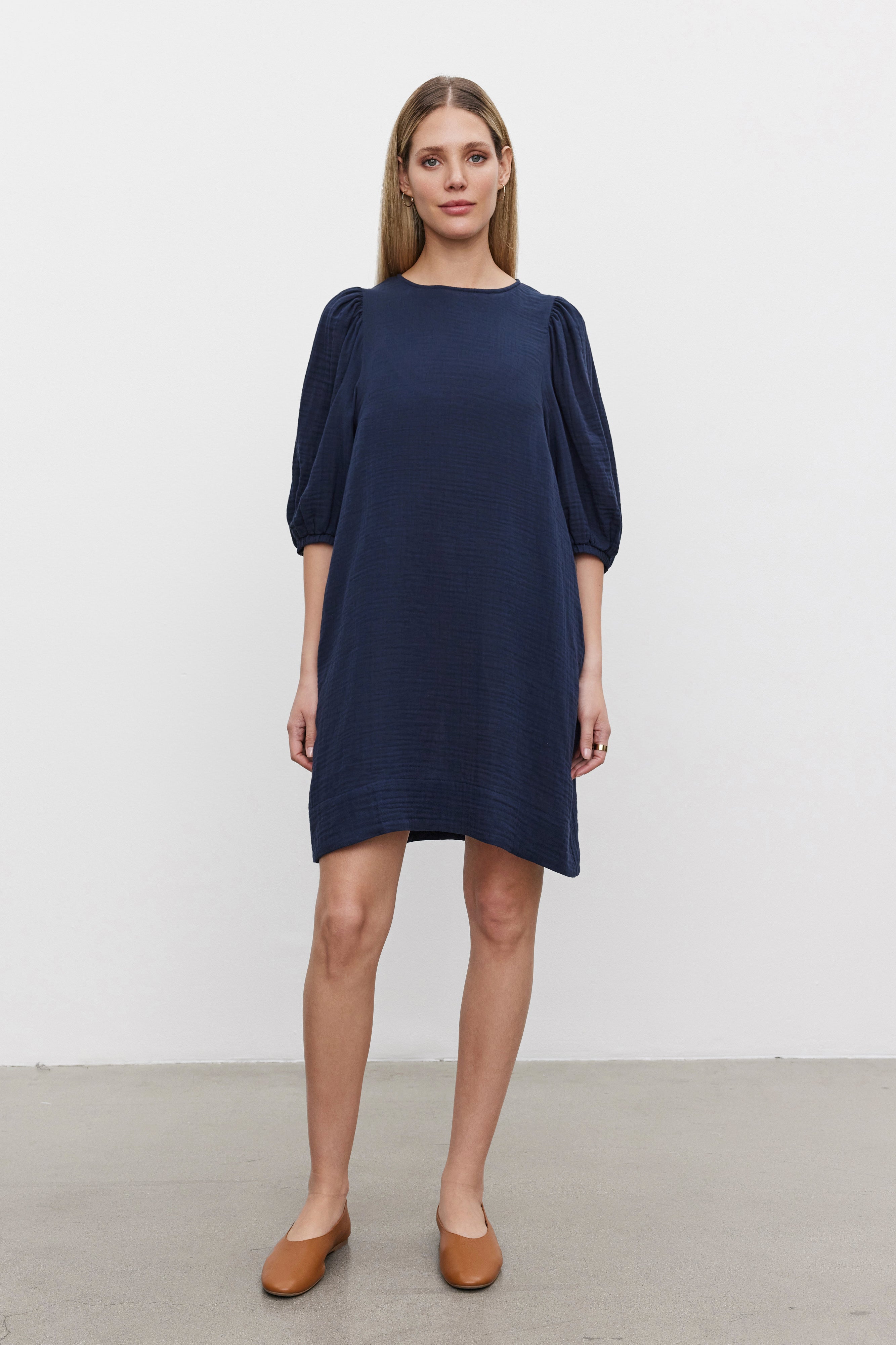   A woman is standing against a plain white background, wearing the KARINA COTTON GAUZE DRESS by Velvet by Graham & Spencer—a navy blue, knee-length dress with puff sleeves—and brown flat shoes, perfect for casual outings. 