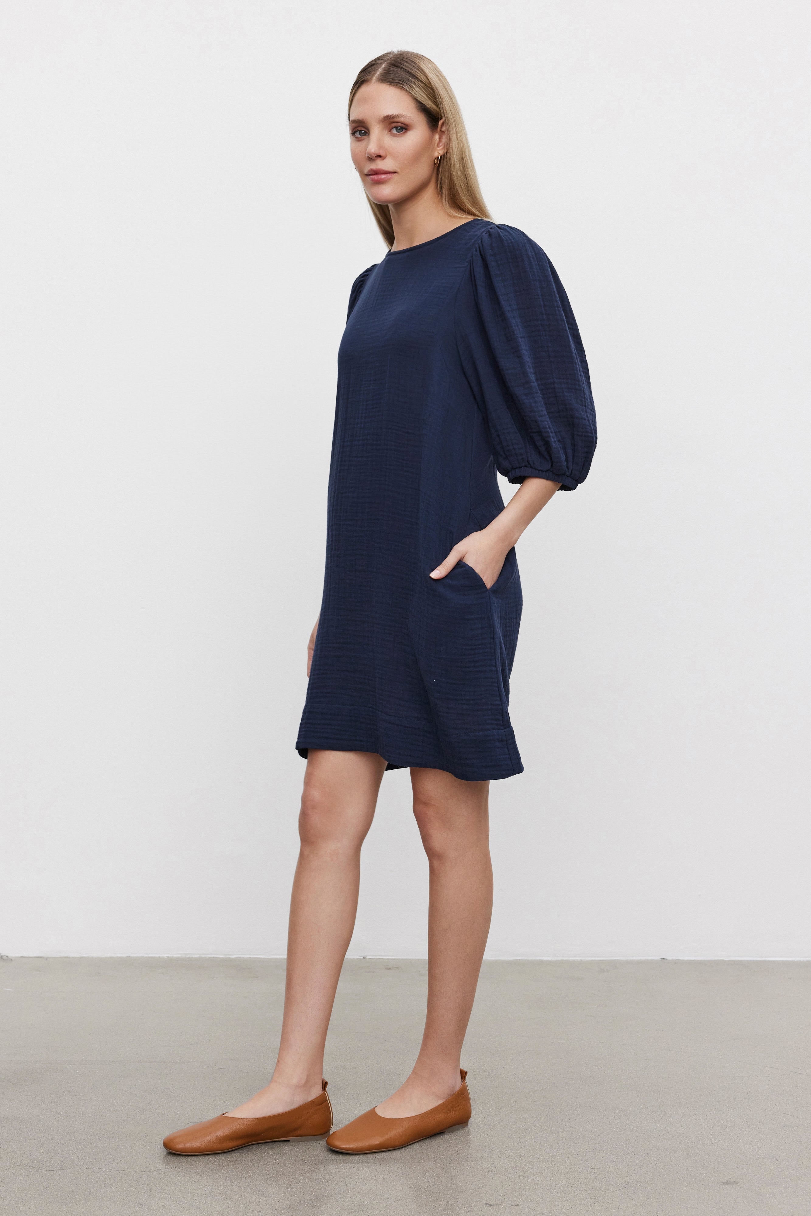   A woman is standing against a plain background, wearing the KARINA Cotton Gauze Dress by Velvet by Graham & Spencer in navy blue with puff sleeves and brown flats. She has one hand in her pocket and is looking towards the camera. 