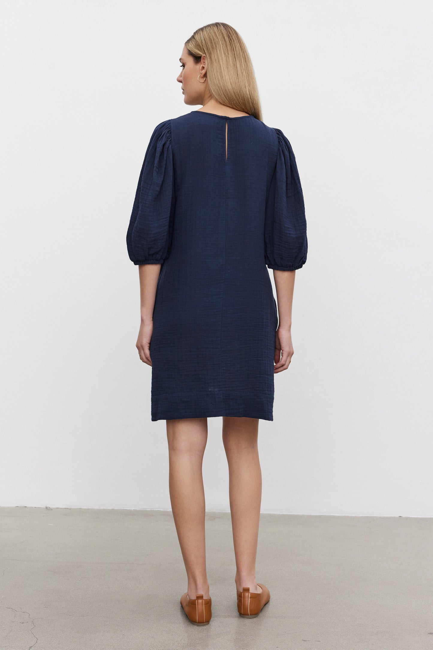 A woman stands facing away, wearing the KARINA COTTON GAUZE DRESS by Velvet by Graham & Spencer. The dark blue, knee-length dress features puff sleeves with elastic cuffs. She has shoulder-length blonde hair and brown loafers on her feet. The background consists of a plain white wall and a grey floor.-37629603086529