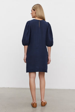 A woman stands facing away, wearing the KARINA COTTON GAUZE DRESS by Velvet by Graham & Spencer. The dark blue, knee-length dress features puff sleeves with elastic cuffs. She has shoulder-length blonde hair and brown loafers on her feet. The background consists of a plain white wall and a grey floor.