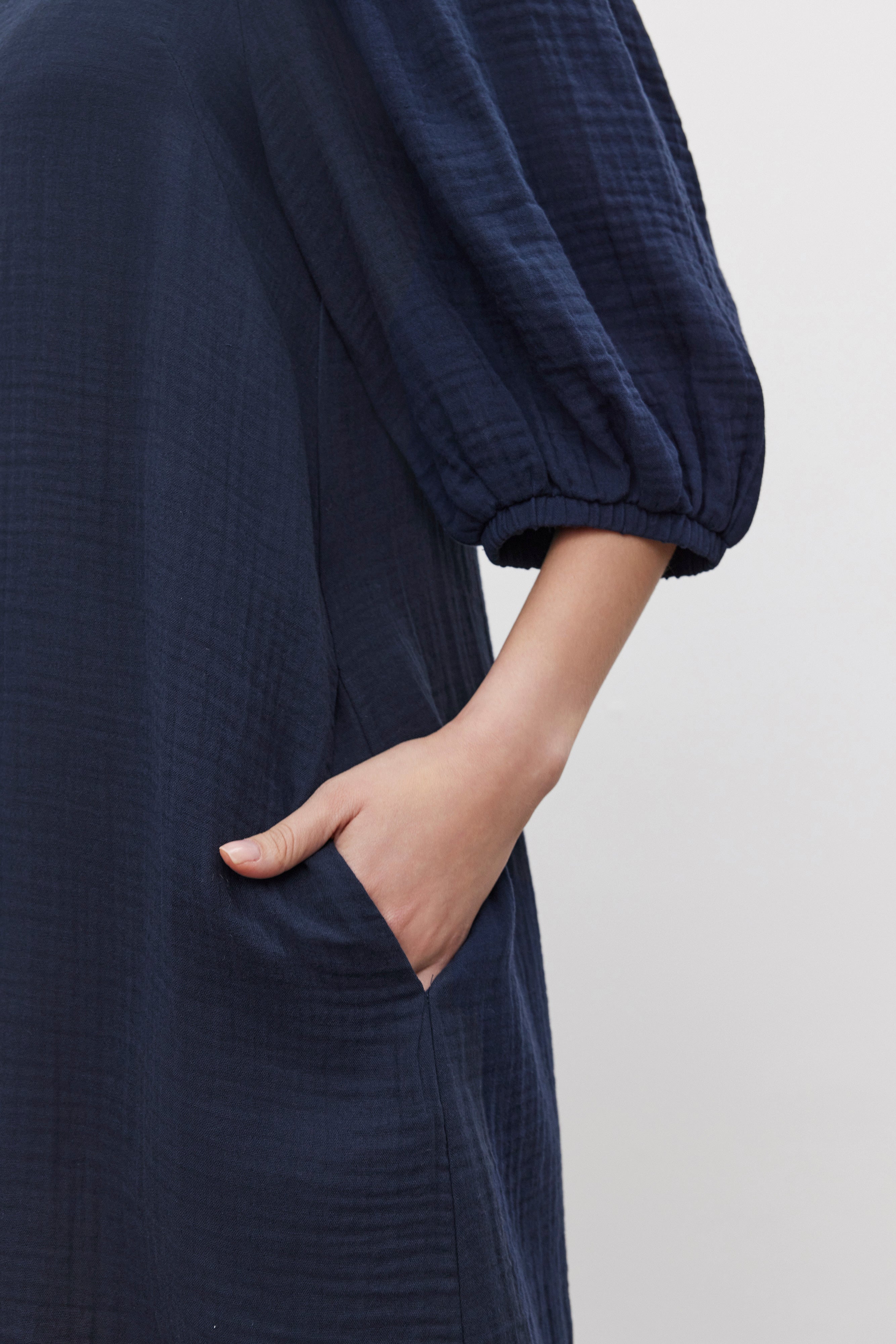   A person donning the KARINA COTTON GAUZE DRESS by Velvet by Graham & Spencer in dark blue, complete with puff sleeves and a convenient pocket, perfect for casual outings. 