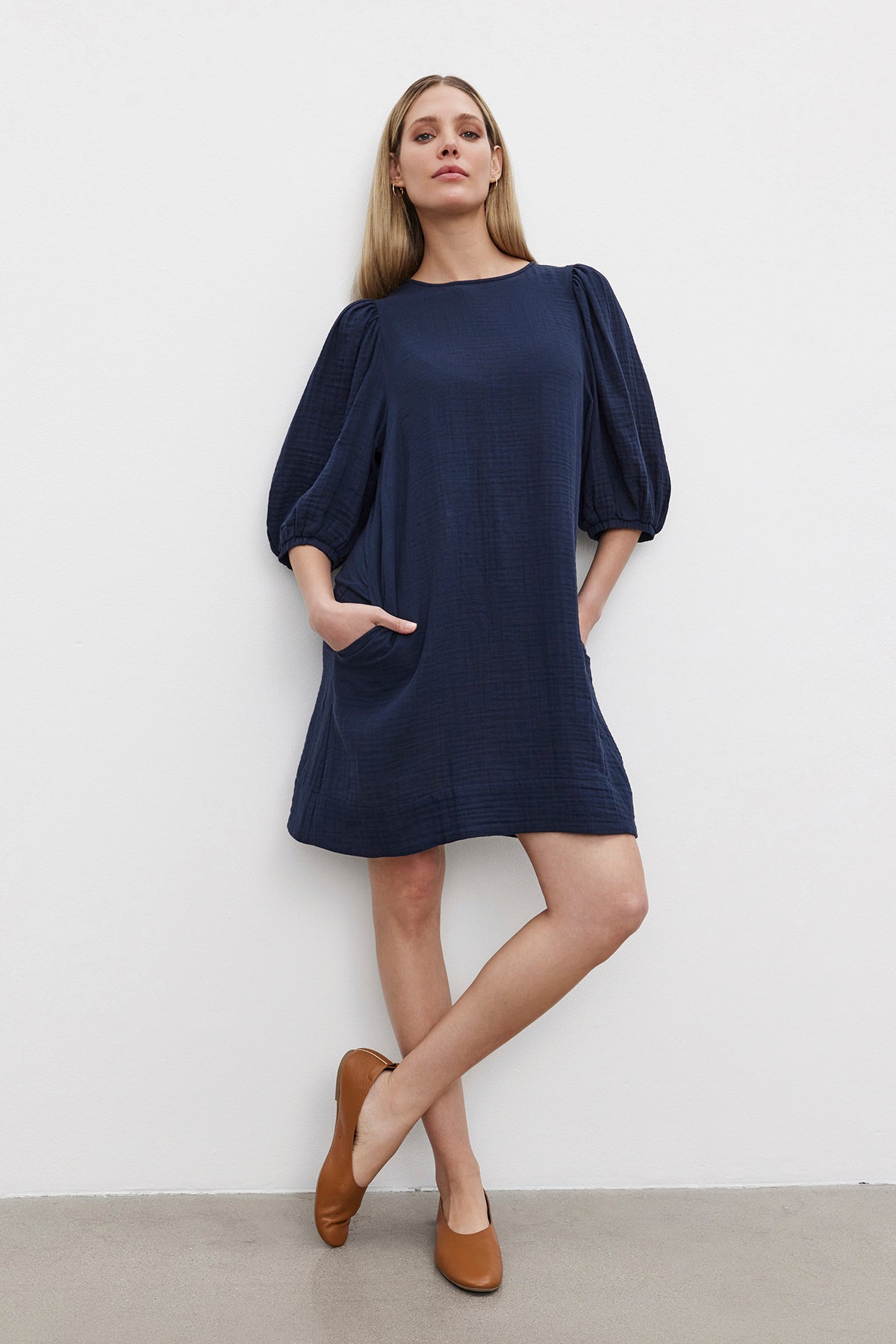   A woman stands against a white wall, wearing the loose-fitting KARINA COTTON GAUZE DRESS by Velvet by Graham & Spencer in navy blue with balloon sleeves and brown flat shoes, her hands tucked in the dress pockets. 