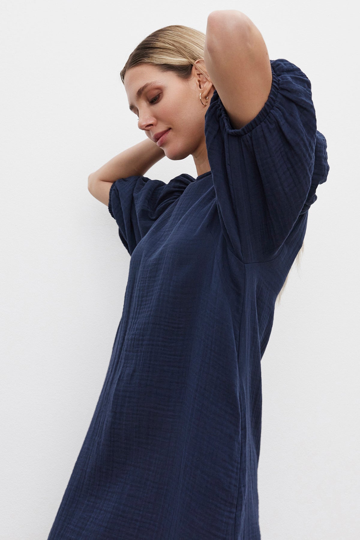   A woman wearing the KARINA COTTON GAUZE DRESS by Velvet by Graham & Spencer, a loose-fitting navy blue dress with elastic cuffs, fixes her hair against a plain white background. 