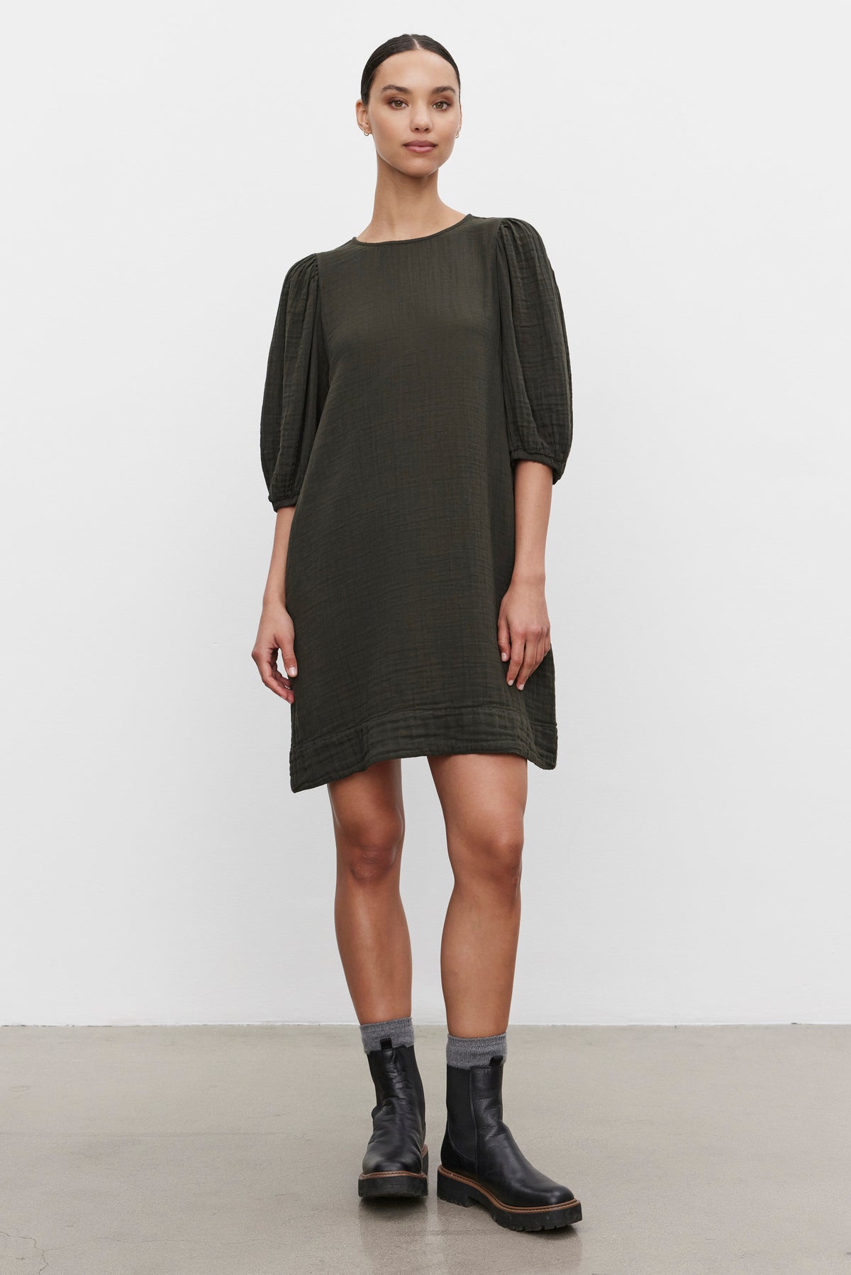   A woman stands against a white background, wearing the KARINA COTTON GAUZE DRESS in dark green from Velvet by Graham & Spencer, which features knee-length design and puff sleeves, paired with black boots. 