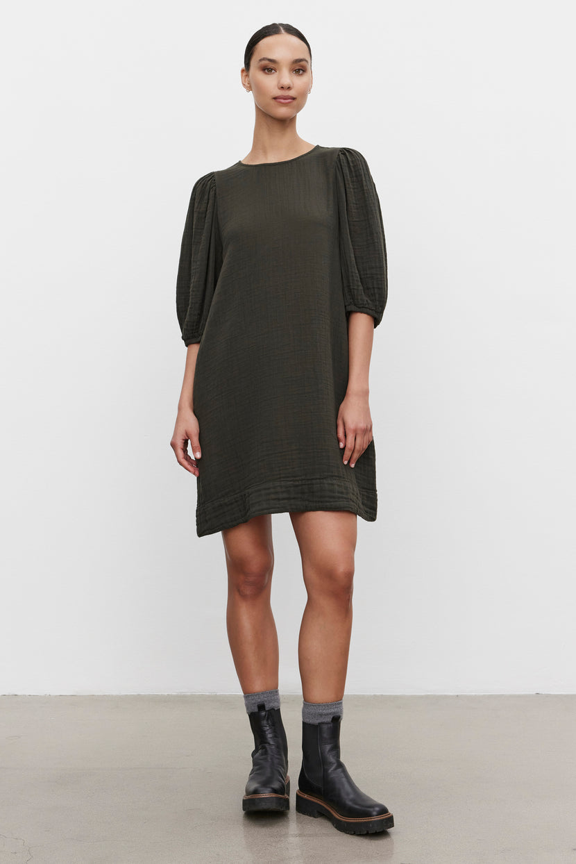 A woman stands against a white background, wearing the KARINA COTTON GAUZE DRESS in dark green from Velvet by Graham & Spencer, which features knee-length design and puff sleeves, paired with black boots.