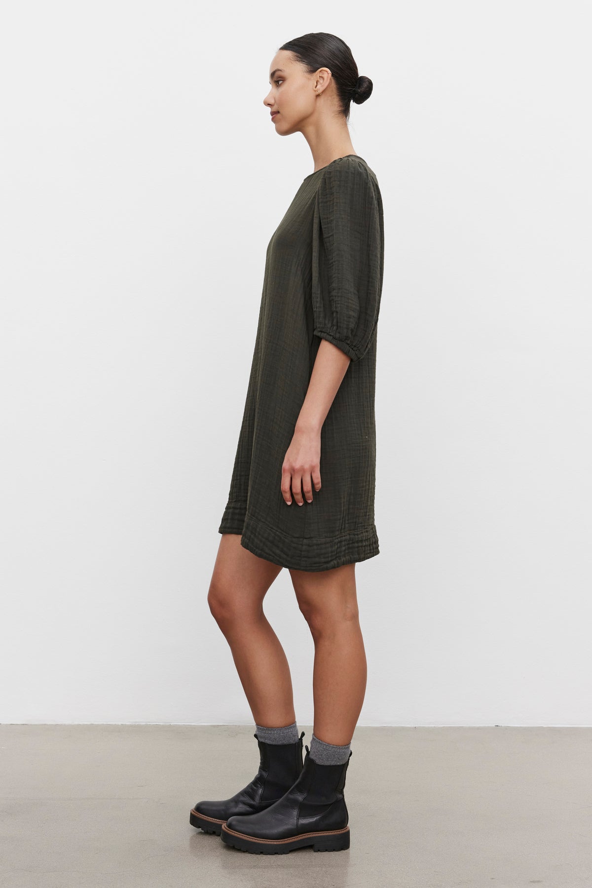   A woman stands sideways wearing a knee-length KARINA COTTON GAUZE DRESS in dark green from Velvet by Graham & Spencer, along with black ankle boots and gray socks, against a plain white background. 