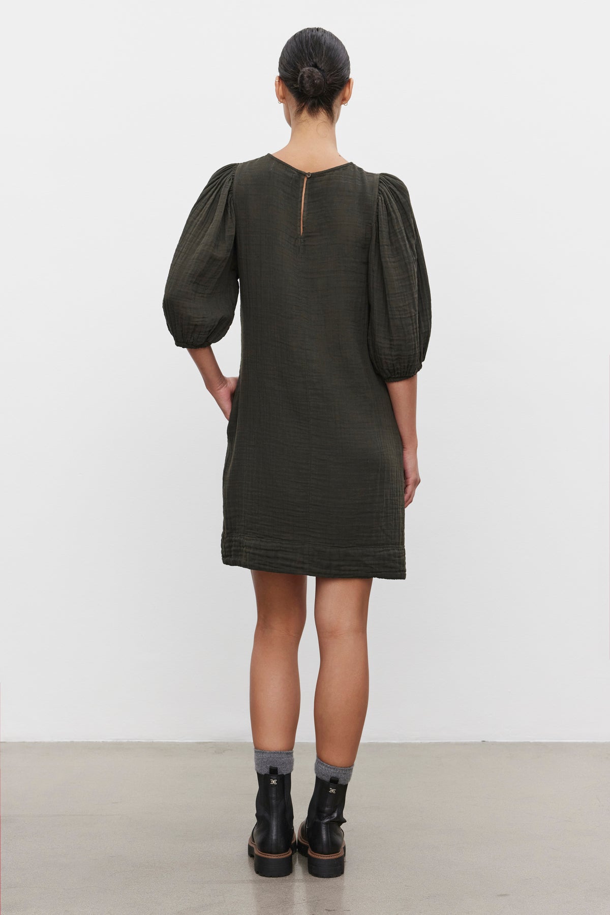   A person with their hair tied back stands with their back to the camera, wearing a dark green KARINA COTTON GAUZE DRESS from Velvet by Graham & Spencer, paired with black ankle boots. 