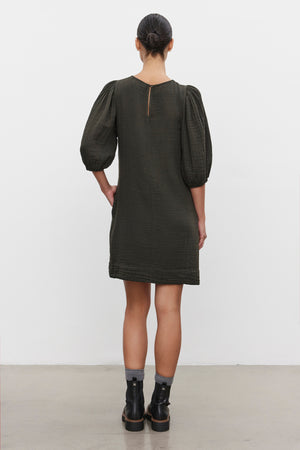 A person with their hair tied back stands with their back to the camera, wearing a dark green KARINA COTTON GAUZE DRESS from Velvet by Graham & Spencer, paired with black ankle boots.