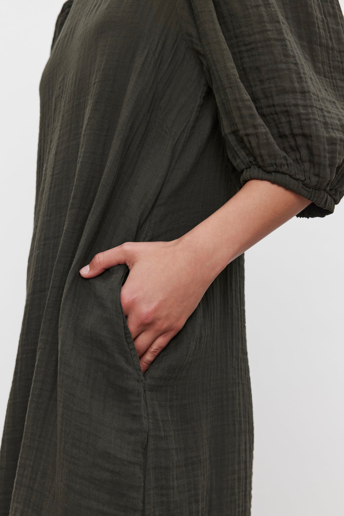   Close-up of a person wearing the KARINA COTTON GAUZE DRESS by Velvet by Graham & Spencer. The long-sleeve, dark green dress features textured fabric and puff sleeves, with their right hand casually placed in the dress pocket. 